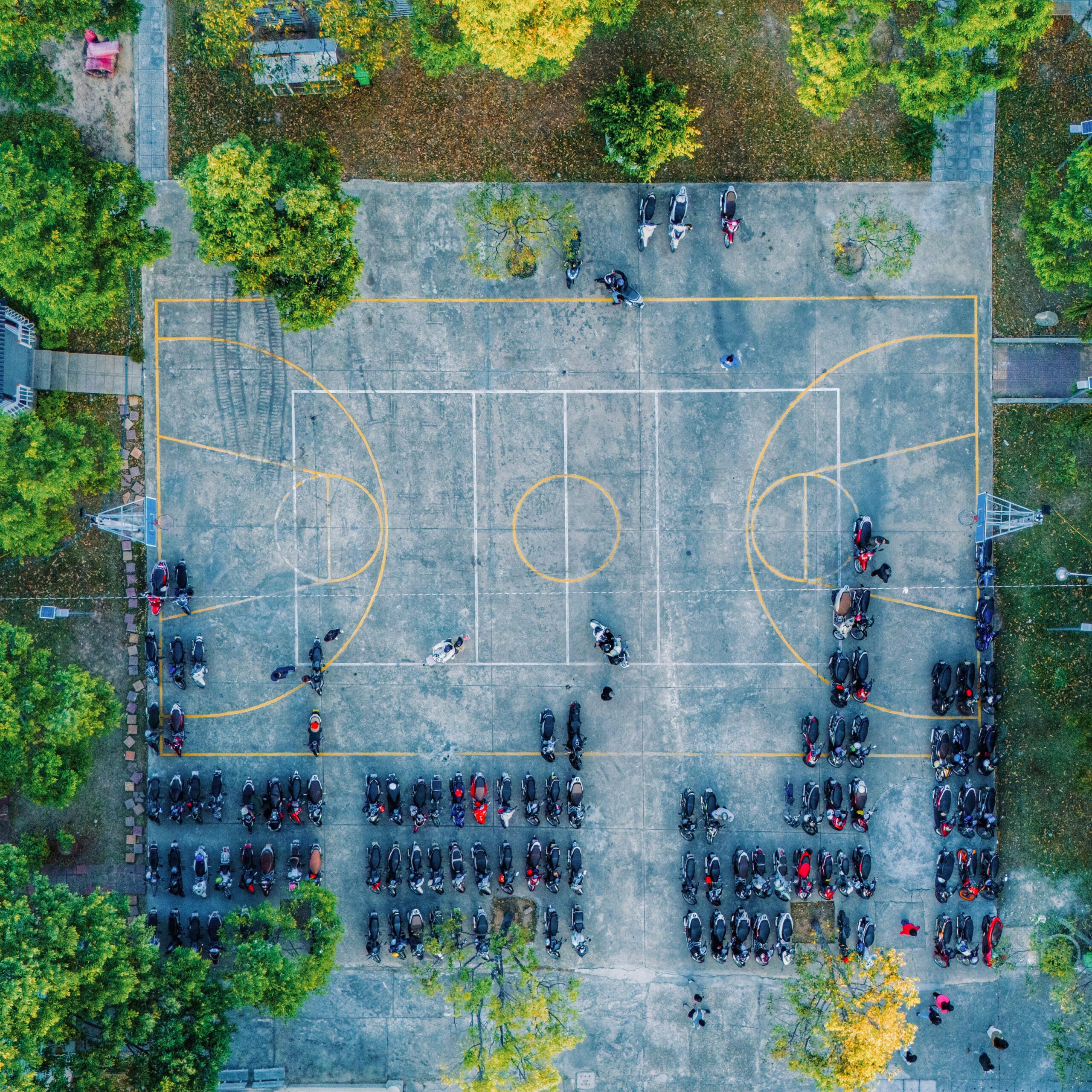 Aerial view of a basketball court surrounded by trees and parked motorbikes.